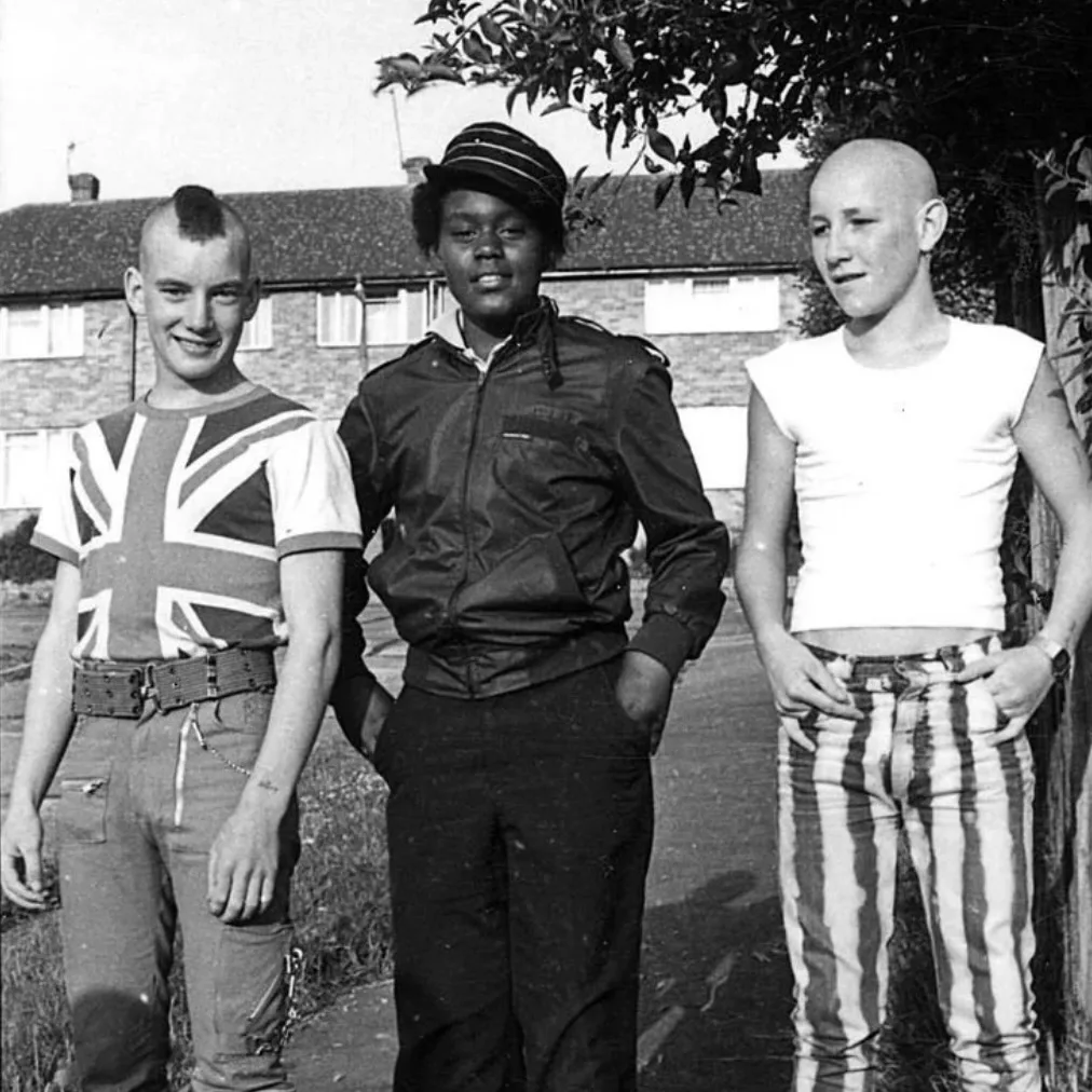 Photo en noir et blanc de trois jeunes posant ensemble devant un immeuble de logements sociaux, l'un en skinhead, l'autre en style rude boy, le troisième avec une crête et un t-shirt Union Jack