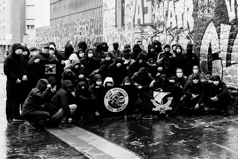 Photo de groupe en noir et blanc d'un cortège antifasciste, visages masqués, banderoles Action Antifasciste Paris Banlieue et Paris Antifa devant un mur recouvert de graffitis
